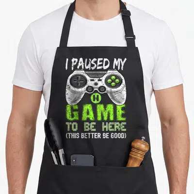 Cute paused video game apron - Distressed game controller with I paused my game text and lime green accents — shown on a black cotton canvas apron against a white background