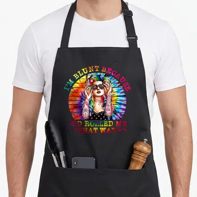 Blunt hippie apron - Rainbow text I'M BLUNT BECAUSE GOD ROLLED ME THAT WAY around tattooed woman with heart sunglasses — shown on a black cotton canvas apron against a white background