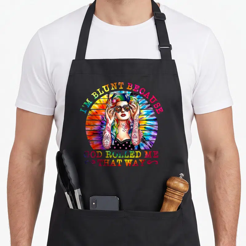 Blunt hippie apron - Rainbow text I'M BLUNT BECAUSE GOD ROLLED ME THAT WAY around tattooed woman with heart sunglasses — shown on a black cotton canvas apron against a white background