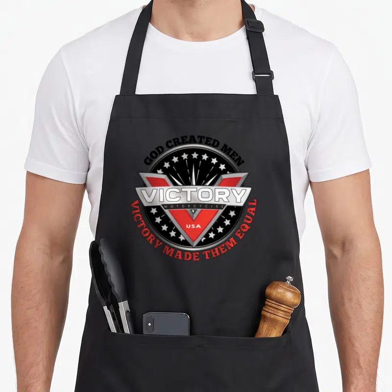 God motorcycle apron - Circular logo with red V, silver banner reading VICTORY, stars, and arched text GOD CREATED MEN — shown on a black cotton canvas apron against a white background