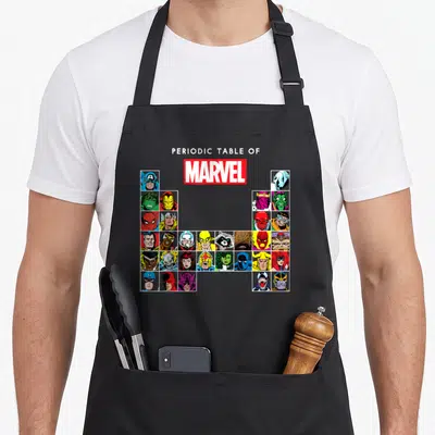 Heroe table apron - Periodic Table of Marvel text above U-grid of cartoon character portraits in primary colors — shown on a black cotton canvas apron against a white background