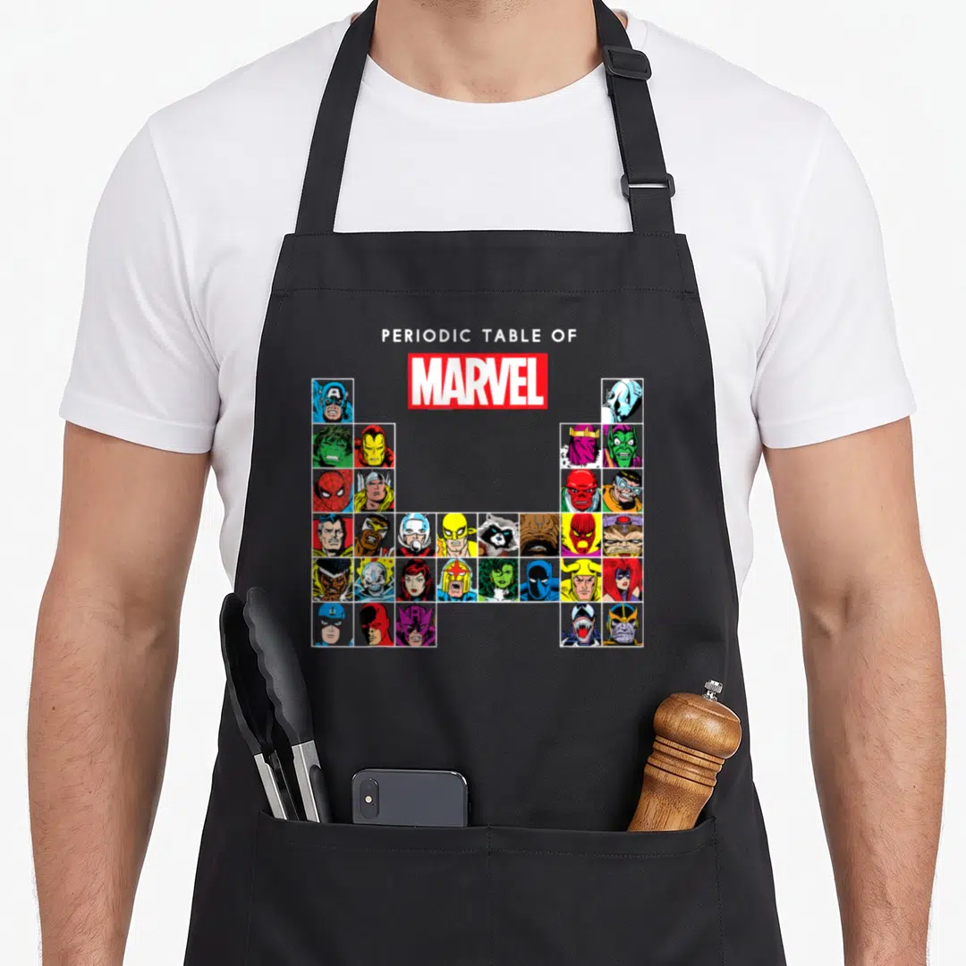 Heroe table apron - Periodic Table of Marvel text above U-grid of cartoon character portraits in primary colors — shown on a black cotton canvas apron against a white background