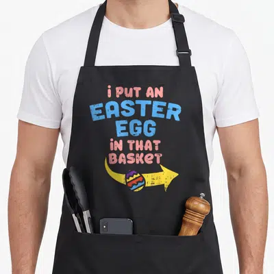 Pregnancy basket apron - i put an easter egg in that basket text with yellow arrow and cracked cartoon egg — shown on a black cotton canvas apron against a white background