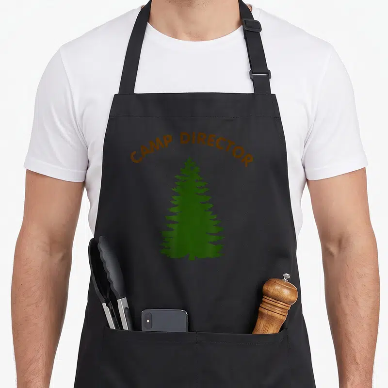 Pine camp chef apron - CAMP DIRECTOR text arched over a green minimalist pine tree silhouette — shown on a black cotton canvas apron against a white background