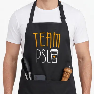 Psl pumpkin apron - Orange TEAM text above white PSL and a coffee cup with an orange heart — shown on a black cotton canvas apron against a white background