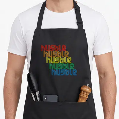 Hustle 70s apron - Stacked red orange yellow green blue hustle typography on black bib — shown on a black cotton canvas apron against a white background