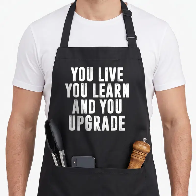 Upgrade divorce party apron - White distressed text reading YOU LIVE YOU LEARN AND YOU UPGRADE stacked vertically — shown on a black cotton canvas apron against a white background