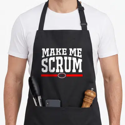 Scrum rugby mom apron - White MAKE ME SCRUM text above red line and white oval — shown on a black cotton canvas apron against a white background