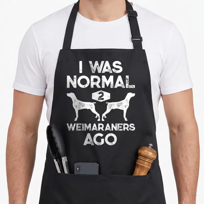Normal weimaraner dog apron - White distressed text I Was Normal 2 Weimaraners Ago above two facing away dog silhouettes — shown on a black cotton canvas apron against a white background