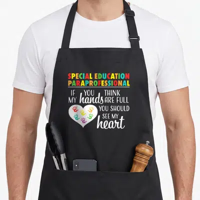 Appreciation education apron - SPECIAL EDUCATION PARAPROFESSIONAL text with colorful handprint heart — shown on a black cotton canvas apron against a white background