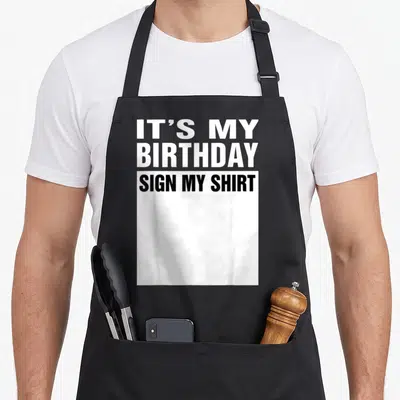 Funny sign birthday apron - Stacked IT'S MY BIRTHDAY SIGN MY SHIRT text above a white rectangle — shown on a black cotton canvas apron against a white background