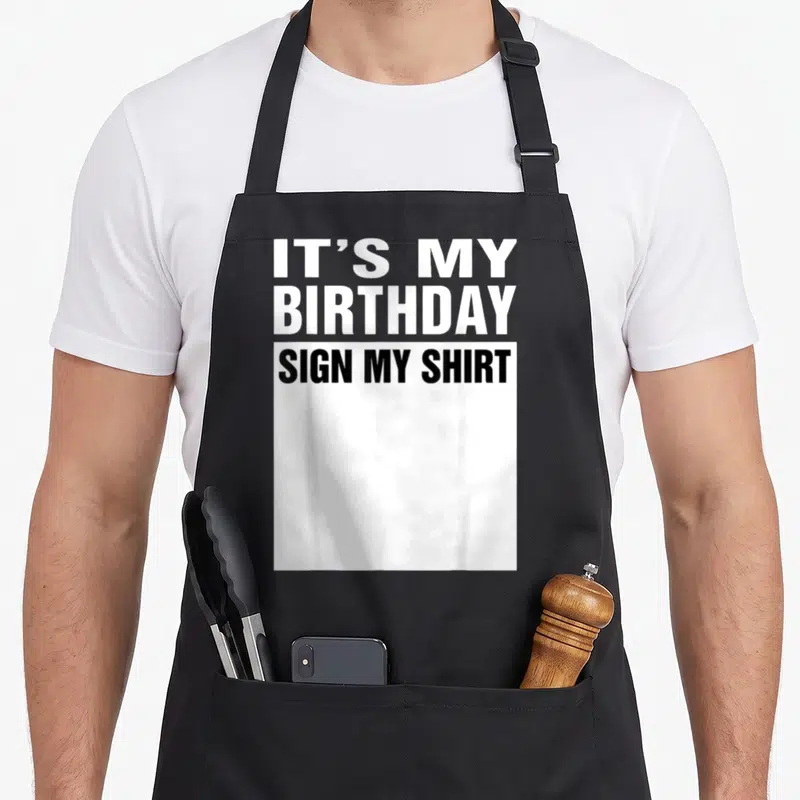 Funny sign birthday apron - Stacked IT'S MY BIRTHDAY SIGN MY SHIRT text above a white rectangle — shown on a black cotton canvas apron against a white background