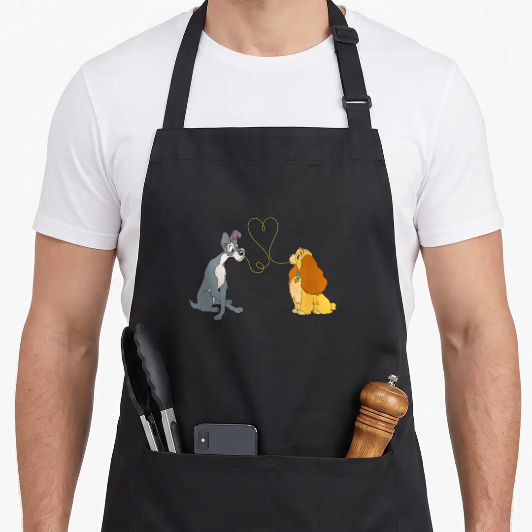 Bella notte lady and the tramp apron - Two cartoon dogs face each other with yellow leash forming heart shape between — shown on a black cotton canvas apron against a white background