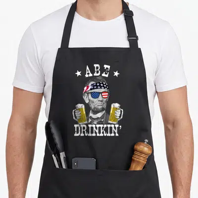 Abe patriotic apron - Abraham Lincoln portrait with flag bandana, sunglasses, and two beer mugs surrounded — shown on a black cotton canvas apron against a white background