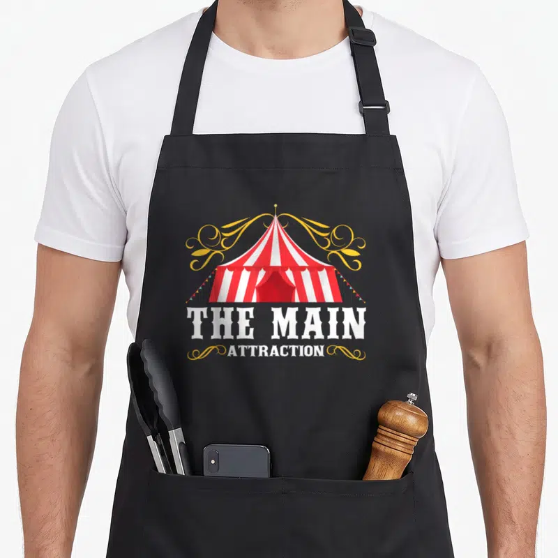 Attraction birthday apron - Red and white striped circus tent with yellow swirls and stacked white text reading — shown on a black cotton canvas apron against a white background