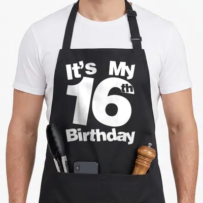Vintage 16th birthday apron - White text It's My 16th Birthday stacked vertically with large number 16 — shown on a black cotton canvas apron against a white background