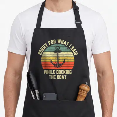 Docking boat apron - Retro striped sun with anchor and text SORRY FOR WHAT I SAID WHILE DOCKING THE BOAT — shown on a black cotton canvas apron against a white background