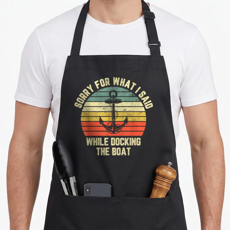 Docking boat apron - Retro striped sun with anchor and text SORRY FOR WHAT I SAID WHILE DOCKING THE BOAT — shown on a black cotton canvas apron against a white background