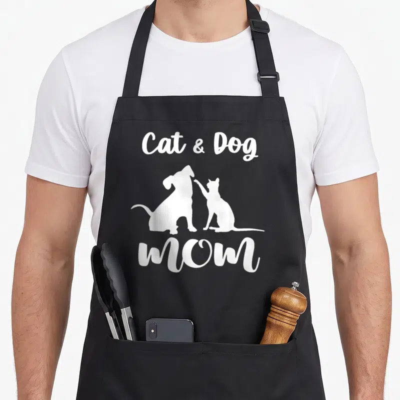 Dog pet cat apron - White Cat & Dog MOM text above and below dog and cat silhouettes touching paws — shown on a black cotton canvas apron against a white background