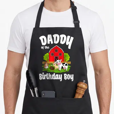 Bday farm aprons - Cartoon red barn with four farm animals and Daddy of the Birthday Boy text — shown on a black cotton canvas apron against a white background