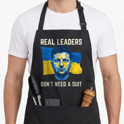 Leader ukrainian aprons - Stylized portrait of Zelenskyy on flag with text Real Leaders Don't Need A Suit — shown on a black cotton canvas apron against a white background