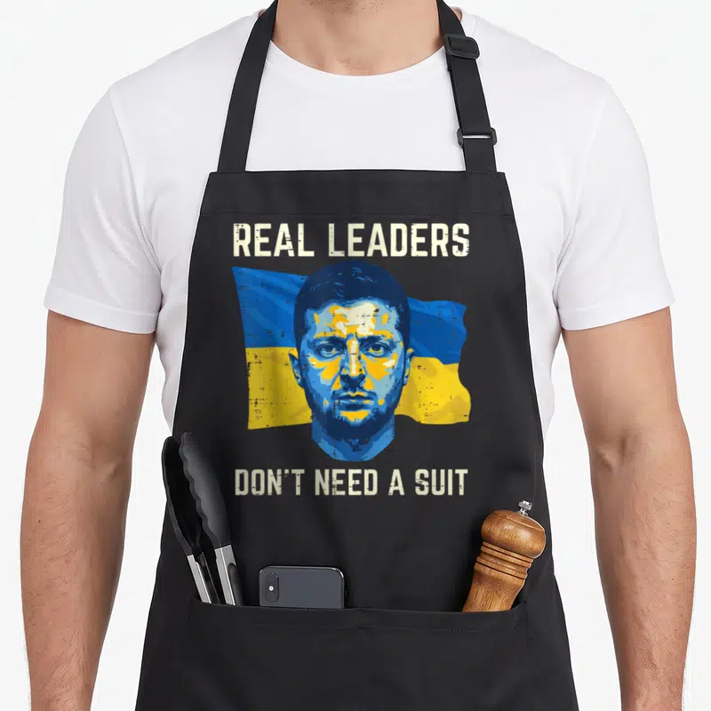 Leader ukrainian aprons - Stylized portrait of Zelenskyy on flag with text Real Leaders Don't Need A Suit — shown on a black cotton canvas apron against a white background
