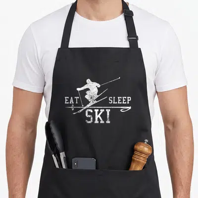 Ski sleep apron - White distressed skier silhouette flanked by EAT SLEEP SKI text — shown on a black cotton canvas apron against a white background