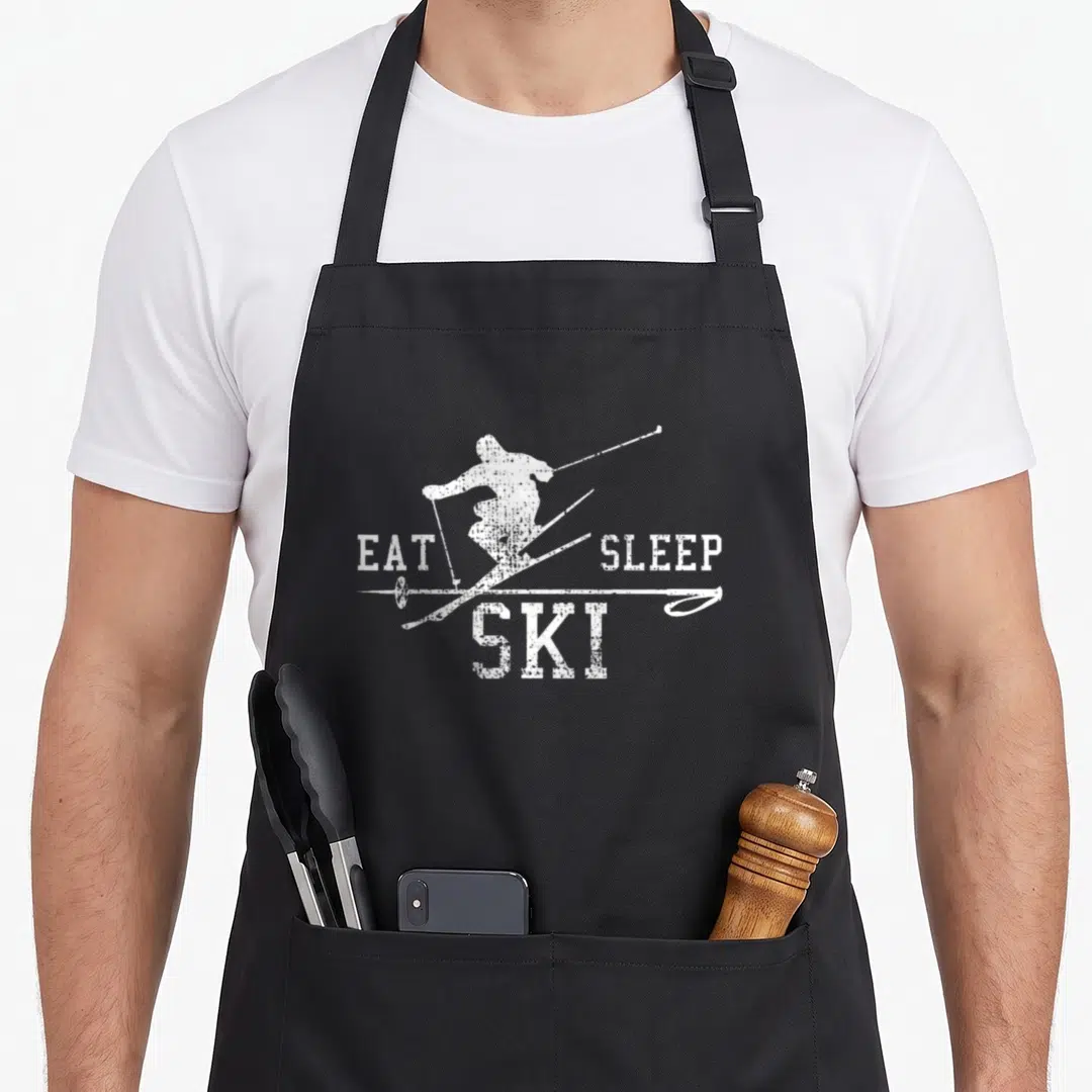 Ski sleep apron - White distressed skier silhouette flanked by EAT SLEEP SKI text — shown on a black cotton canvas apron against a white background
