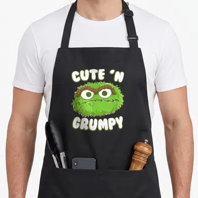 Grumpy oscar the grouch apron - Green monster head with brown eyebrows between cute n grumpy text — shown on a black cotton canvas apron against a white background