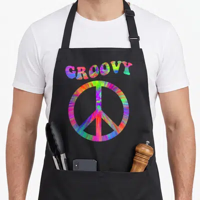 70s tie-dye apron - GROOVY text arched over tie-dye peace symbol in pink blue green yellow orange — shown on a black cotton canvas apron against a white background