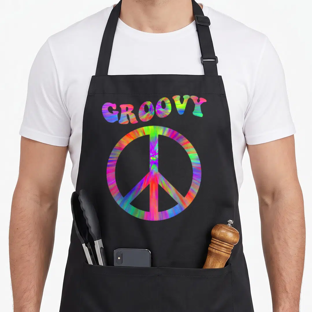 70s tie-dye apron - GROOVY text arched over tie-dye peace symbol in pink blue green yellow orange — shown on a black cotton canvas apron against a white background