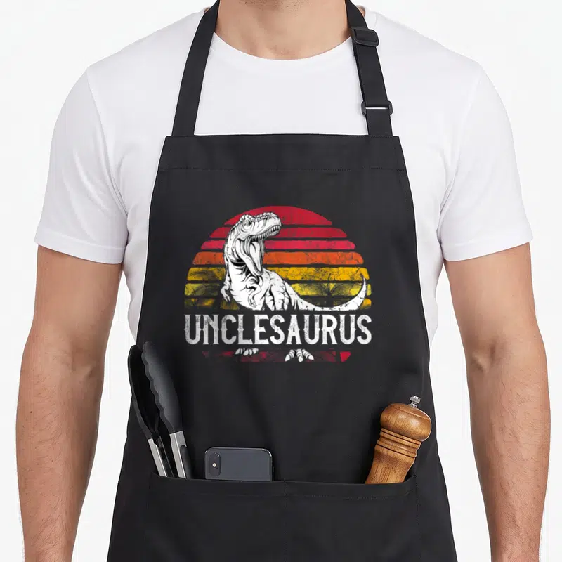 Unclesaurus father day apron - White T-Rex in front of red orange and yellow striped sun with UNCLESAURUS text below — shown on a black cotton canvas apron against a white background