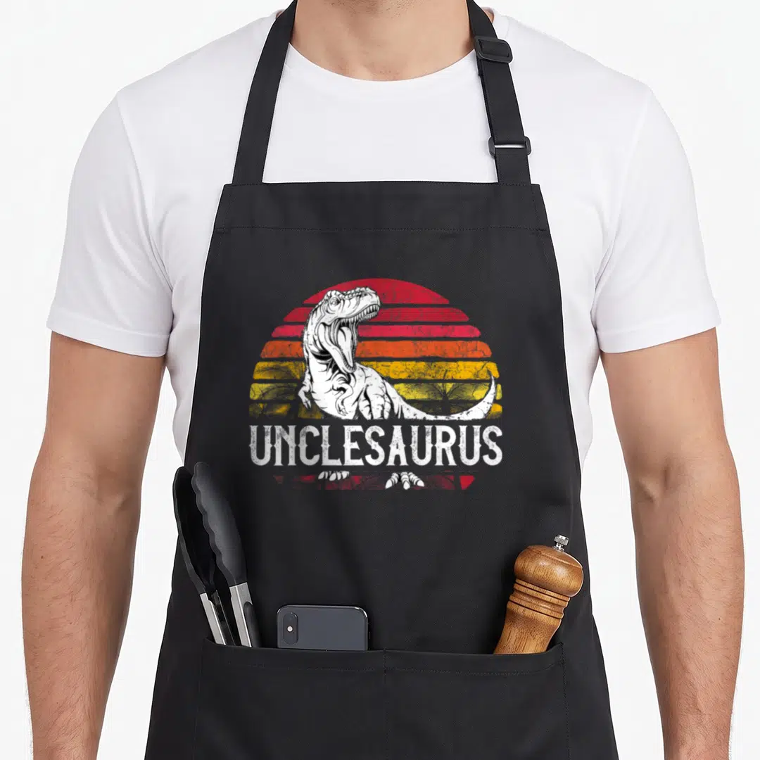 Unclesaurus father day apron - White T-Rex in front of red orange and yellow striped sun with UNCLESAURUS text below — shown on a black cotton canvas apron against a white background
