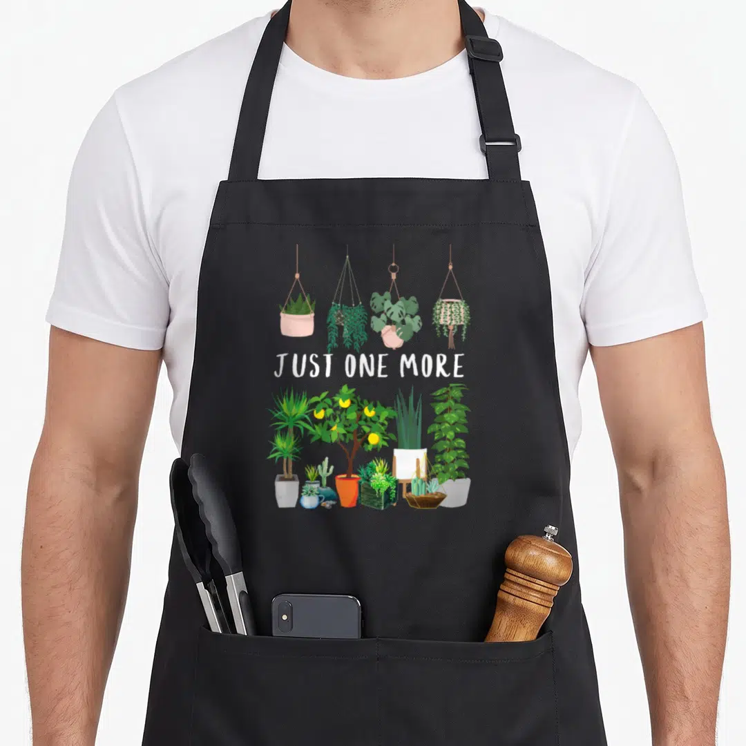 Indoor plant lady apron - JUST ONE MORE text with four hanging plants and various potted houseplants in cartoon style — shown on a black cotton canvas apron against a white background