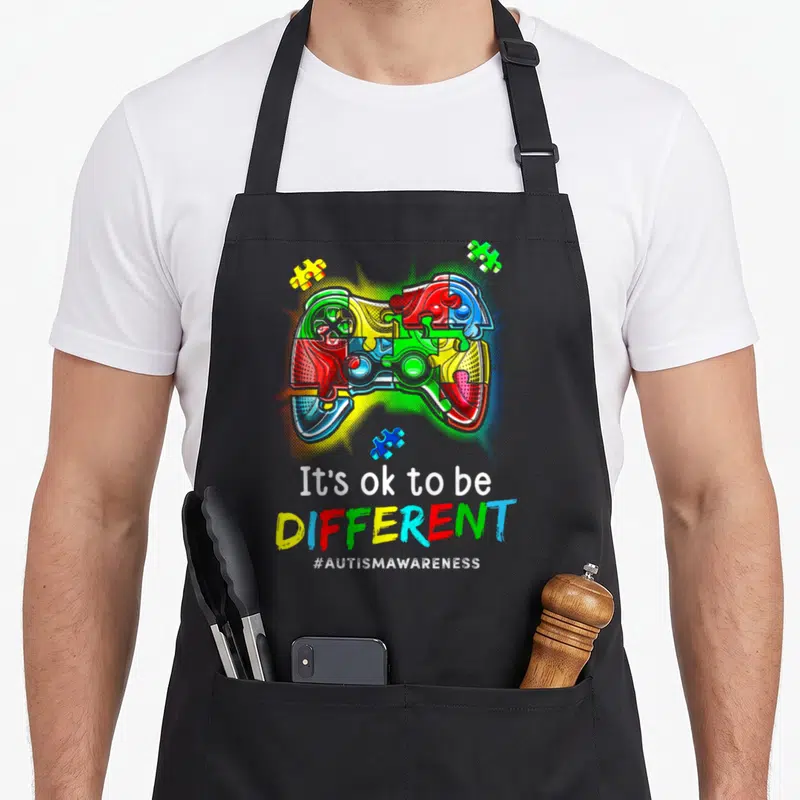 Autism video game apron - Multicolored game controller made of puzzle pieces with It's ok to be DIFFERENT — shown on a black cotton canvas apron against a white background
