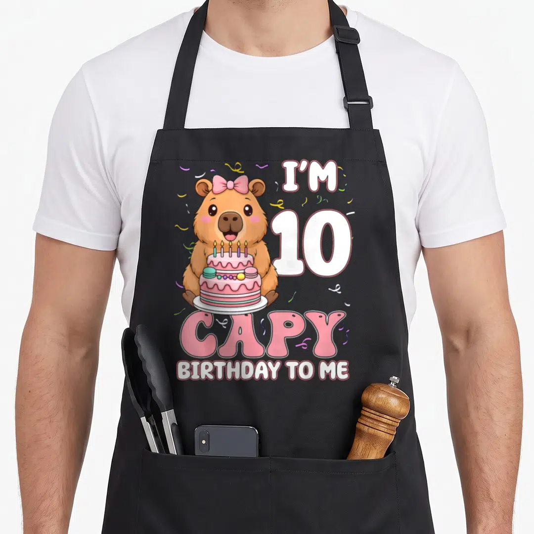 Capy 10th capybara birthday apron - Cartoon brown capybara holding pink cake with I'M 10 CAPY BIRTHDAY TO ME text — shown on a black cotton canvas apron against a white background