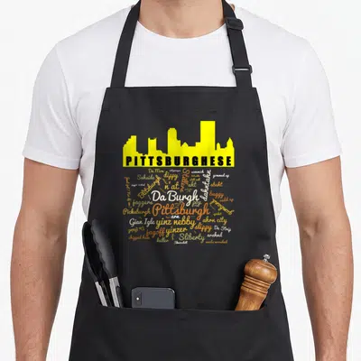 Pittsburghese pittsburgh apron - Yellow Pittsburgh skyline with PITTSBURGHESE text and word cloud of local slang terms — shown on a black cotton canvas apron against a white background