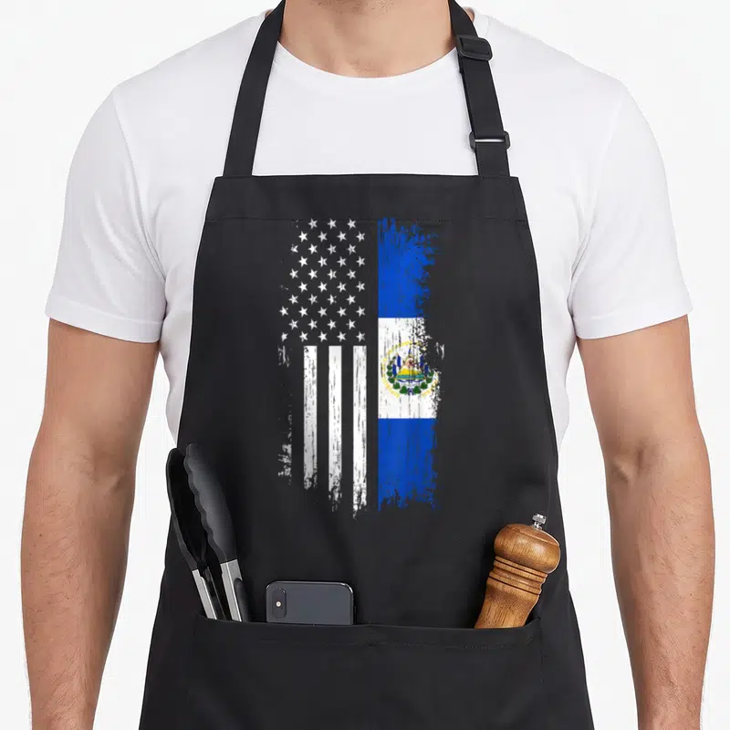 Salvadoran el cook apron - Distressed hybrid flag with USA stars stripes and El Salvador emblem REPUBLICA DE EL SALVADOR — shown on a black cotton canvas apron against a white background