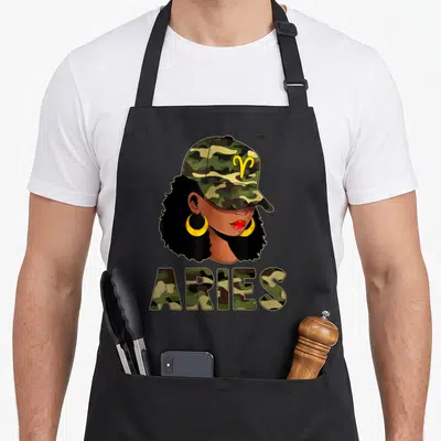 Queen aries kitchen apron - Cartoon woman in camo cap with yellow earrings above ARIES text in green brown black camo — shown on a black cotton canvas apron against a white background