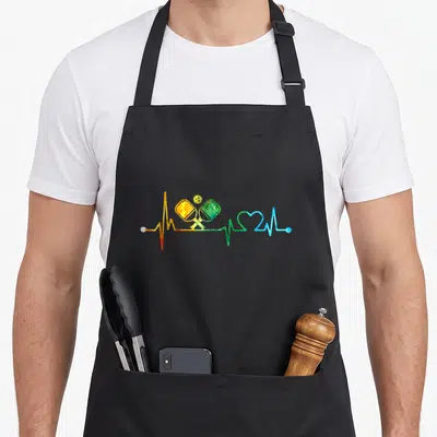 Heartbeat pickleball apron - Rainbow heartbeat line with paddles and ball in red yellow green blue — shown on a black cotton canvas apron against a white background