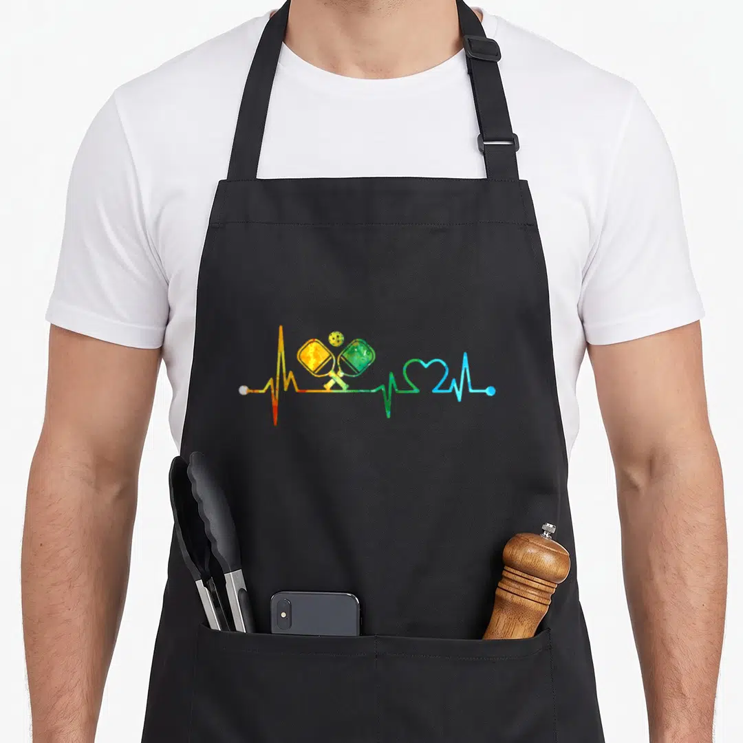 Heartbeat pickleball apron - Rainbow heartbeat line with paddles and ball in red yellow green blue — shown on a black cotton canvas apron against a white background