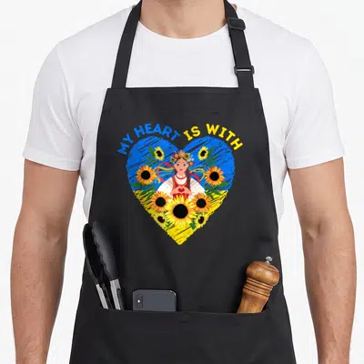 Ukraine traditional apron - Cartoon woman in flower crown inside blue yellow heart with sunflowers and text MY HEART IS — shown on a black cotton canvas apron against a white background