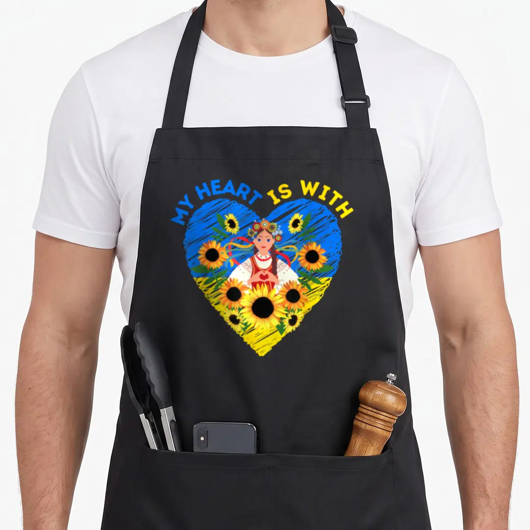 Ukraine traditional apron - Cartoon woman in flower crown inside blue yellow heart with sunflowers and text MY HEART IS — shown on a black cotton canvas apron against a white background