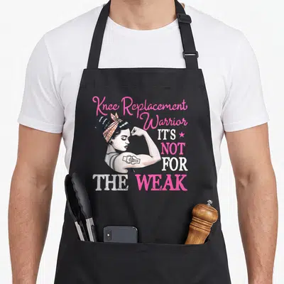 Warrior new knee apron - Cartoon woman flexing arm with bionic tattoo and knee replacement text in pink and white — shown on a black cotton canvas apron against a white background