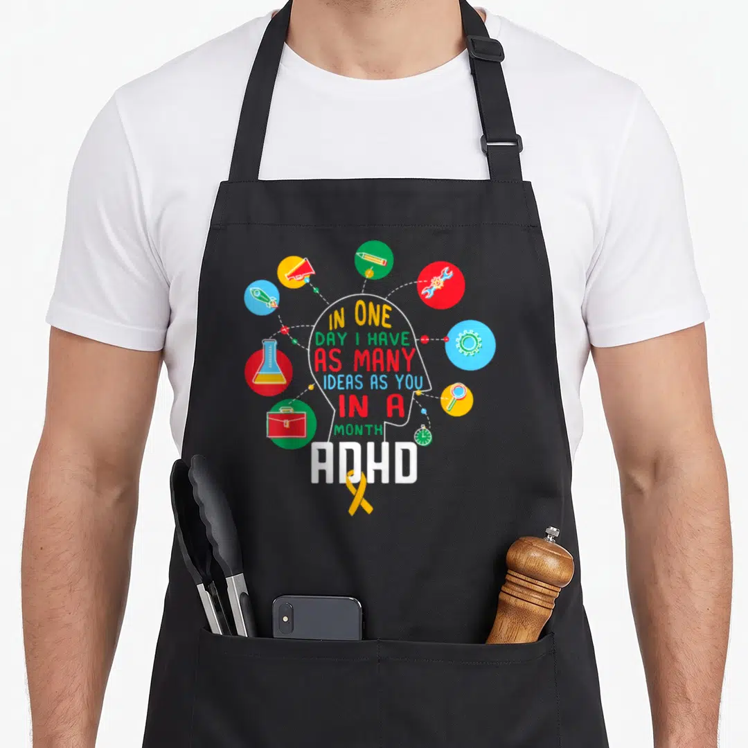 Many adhd awareness apron - White head silhouette with colorful idea circles and ADHD text with yellow ribbon — shown on a black cotton canvas apron against a white background