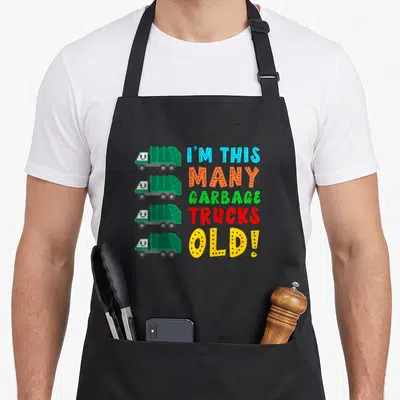 Many garbage truck apron - Four cartoon garbage trucks stacked next to colorful stacked text reading I'M THIS MANY — shown on a black cotton canvas apron against a white background