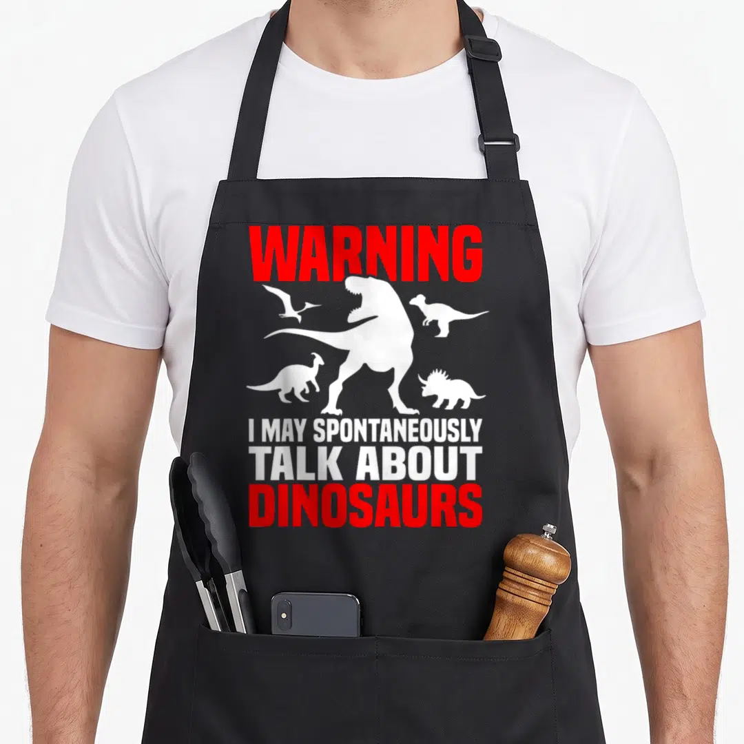 Talking dinosaur apron - Red WARNING text above four white dinosaur silhouettes and three lines of stacked text — shown on a black cotton canvas apron against a white background