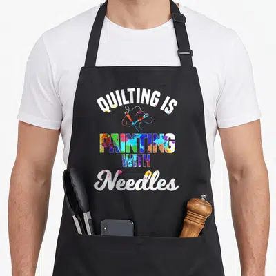 Stitching quilt apron - Stacked text quilting is painting with needles, crossed needles, paint splatters — shown on a black cotton canvas apron against a white background