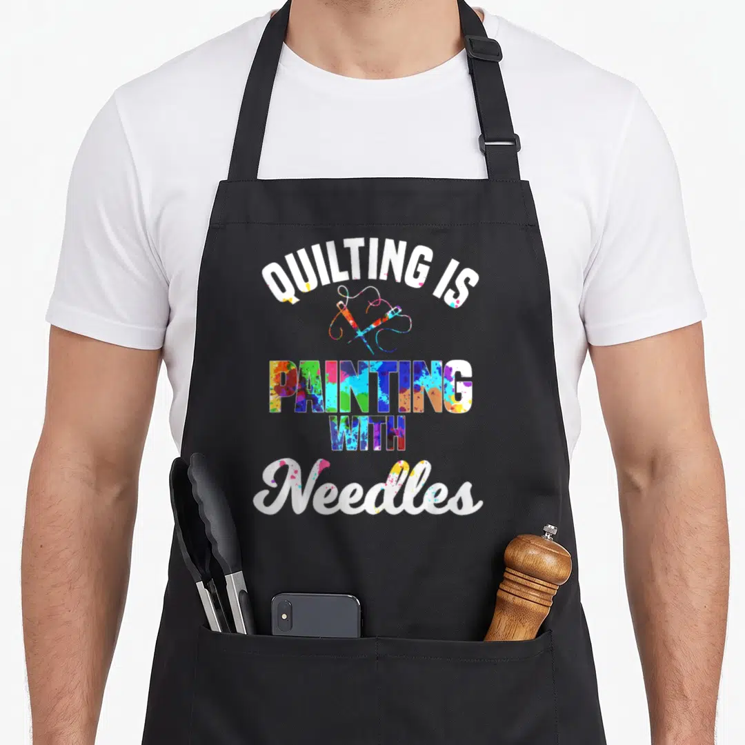 Stitching quilt apron - Stacked text quilting is painting with needles, crossed needles, paint splatters — shown on a black cotton canvas apron against a white background