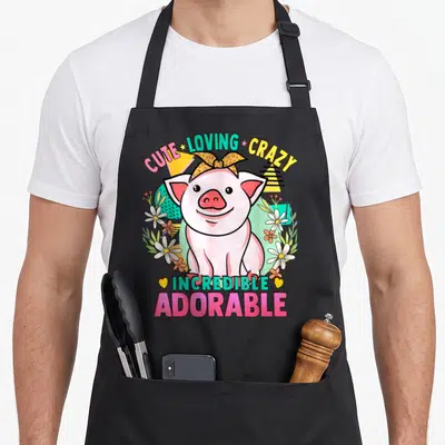 Pig kawaii apron - Cartoon pig with bow and text cute loving crazy incredible adorable surrounded by flowers — shown on a black cotton canvas apron against a white background